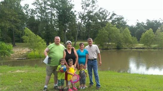 A family stands together by a lake in a park, smiling and enjoying a sunny summer day.