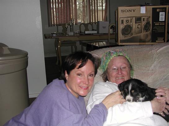Two women smile warmly while holding a small dog, creating a relaxed atmosphere at home.