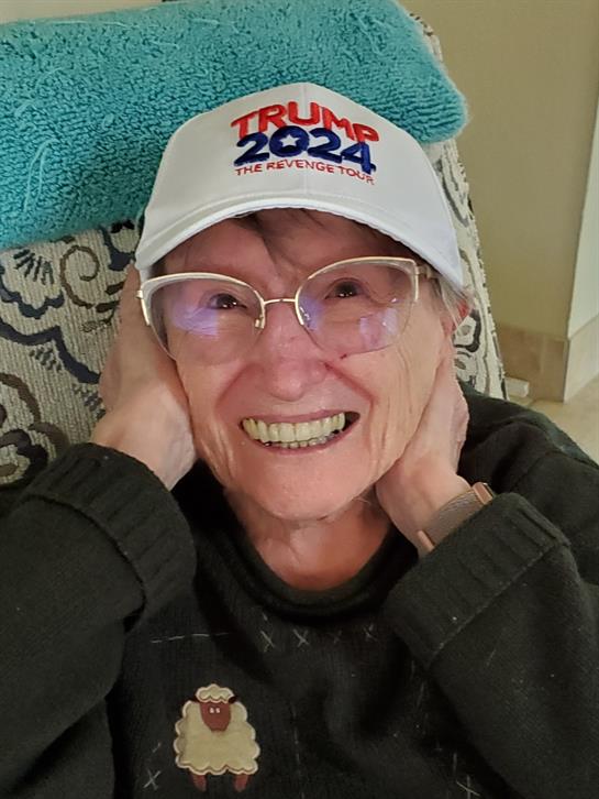 An older woman expresses happiness while sitting indoors, wearing a cap and smiling broadly.