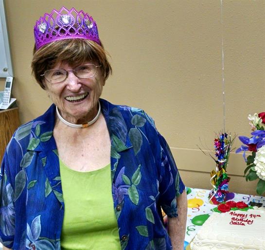 Smiling elderly woman wearing a purple crown enjoys her birthday celebration with a cake nearby.