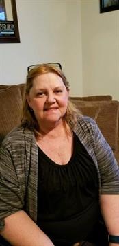 A woman with light brown hair smiles while seated on a couch at home, enjoying a relaxed moment.