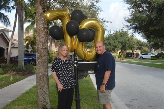 A couple stands by a balloon-decorated mailbox for a 70th birthday in a sunny area.