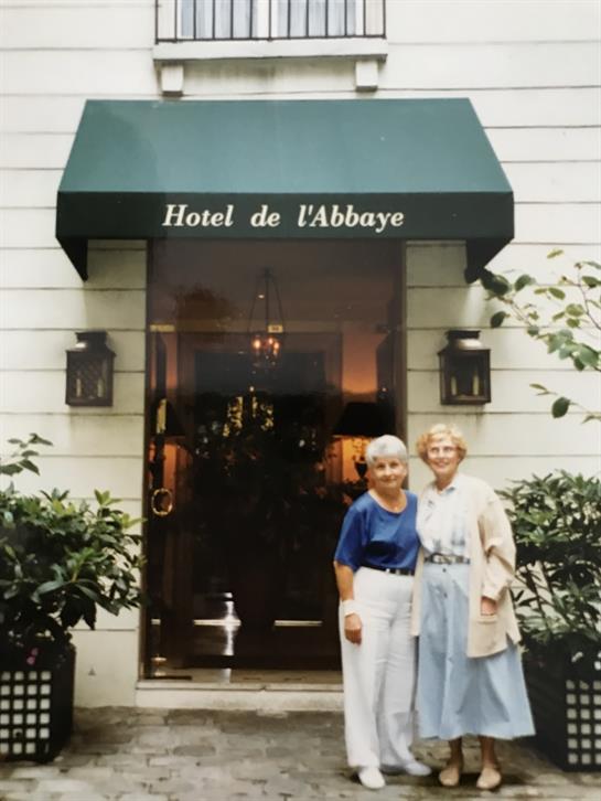 Two elderly women stand outside a hotel, smiling in front of an inviting entrance with plants.