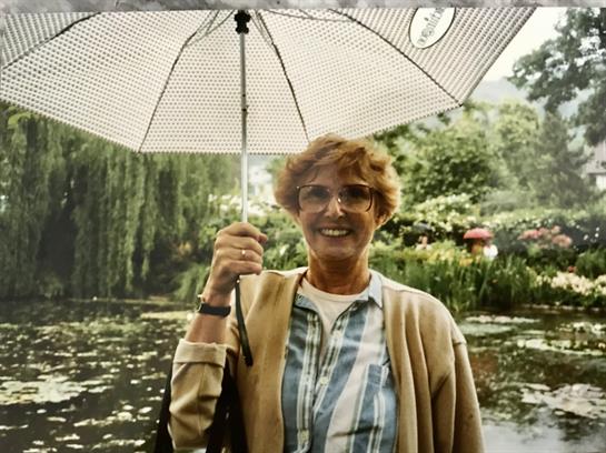 A cheerful older woman stands by a serene pond, holding a white umbrella, surrounded by greenery.