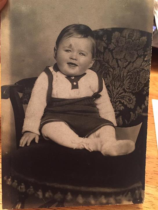 A baby with short hair sits comfortably on a chair, looking cheerful and engaged.
