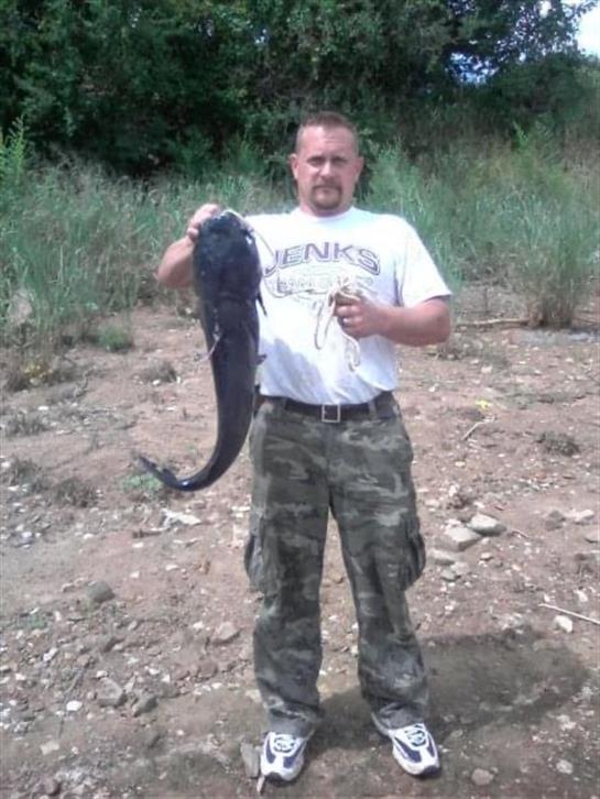 A man proudly displays a large catfish caught from a riverbank, surrounded by greenery.