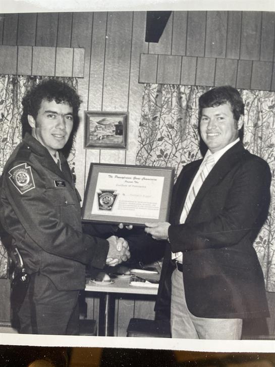 Two men shake hands as one presents a framed certificate to the other in a formal setting.