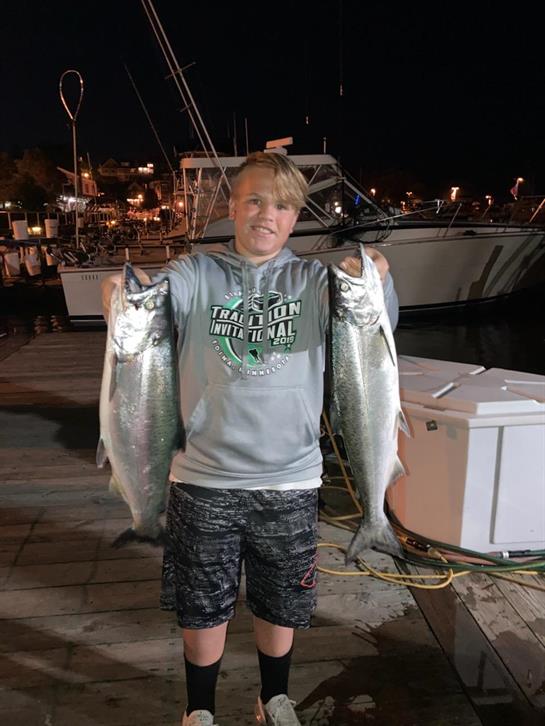A young boy stands at a marina, showcasing two large fish he caught at night.