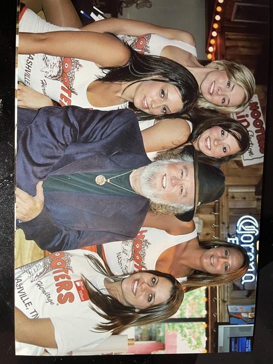 Six women in matching uniforms gather for a cheerful photo with a suited man in a restaurant.