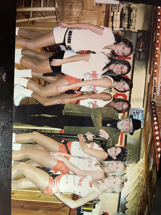 A group of women in matching outfits pose with two men at a lively venue filled with bright lights.