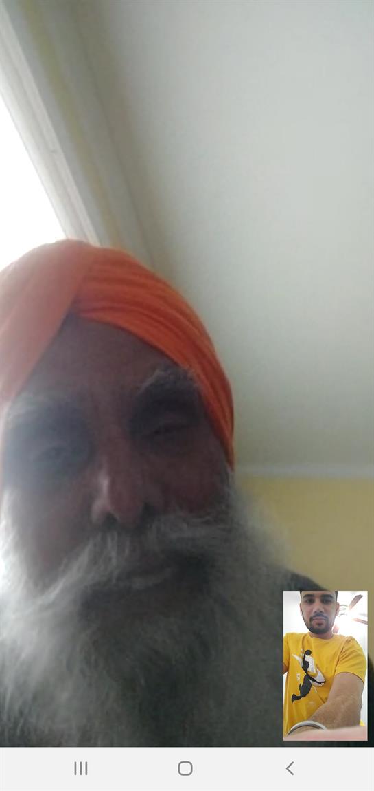 An elderly man wearing an orange turban smiles warmly while sitting indoors during the day.