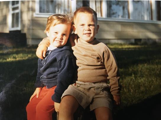 Two boys sit close together, smiling in the afternoon sun with a house backdrop.