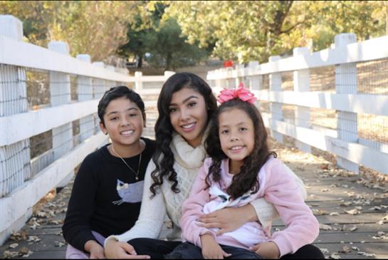 Three children smile while sitting on the ground, enjoying a warm day in a park setting.