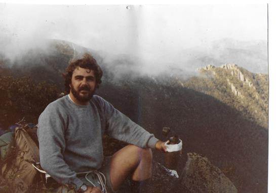 A mountaineer sits on a rocky ledge, sipping from a cup, surrounded by fog and mountain scenery.