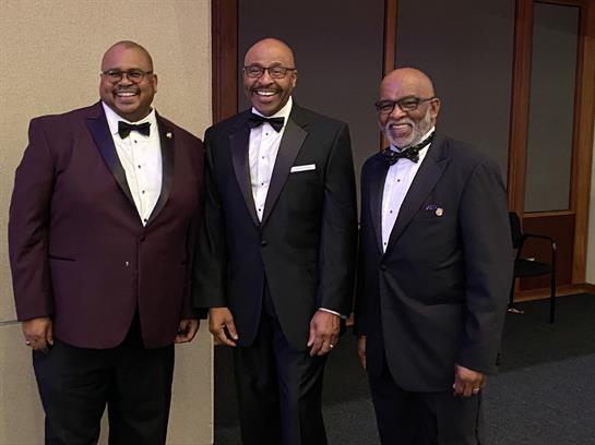 Three men in tuxedos stand together, smiling at a formal gathering during the evening.