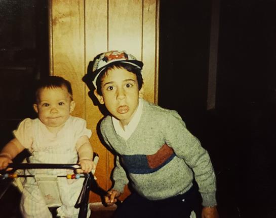 Two young siblings express surprise while standing in their home near a baby stroller.