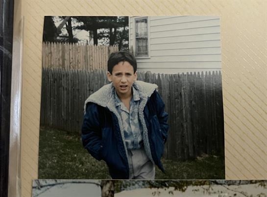 A boy in a jacket poses with a serious expression in front of a wooden fence.
