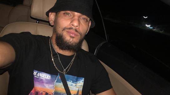 A man with facial hair wears a black bucket hat and graphic t-shirt while sitting in a car at night.