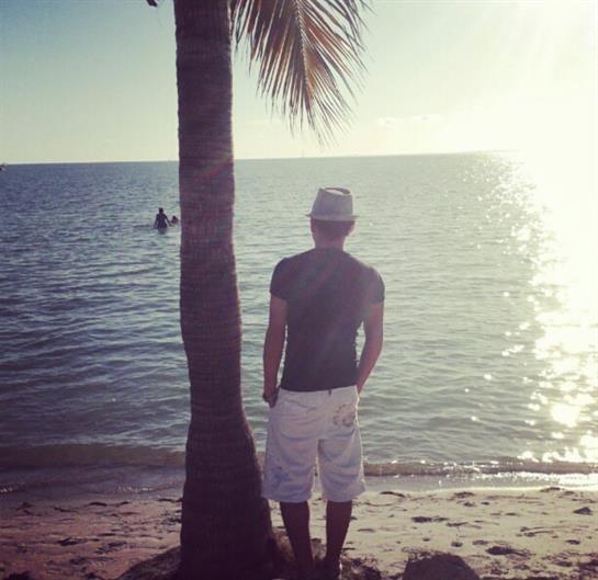 Individual wearing shorts stands near a palm tree at the beach during sunset by the ocean.
