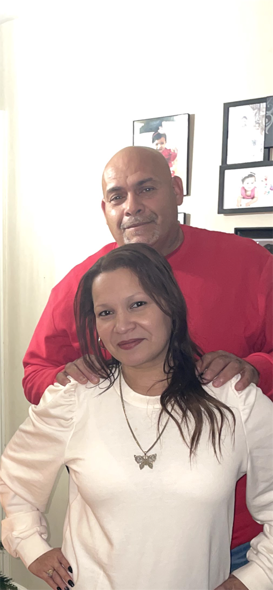 A couple smiles for a picture at their residence, surrounded by framed family memories.