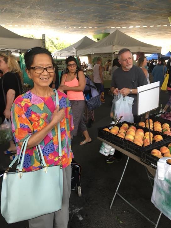 A woman in a colorful outfit joyfully stands at a market with shoppers and baked goods.