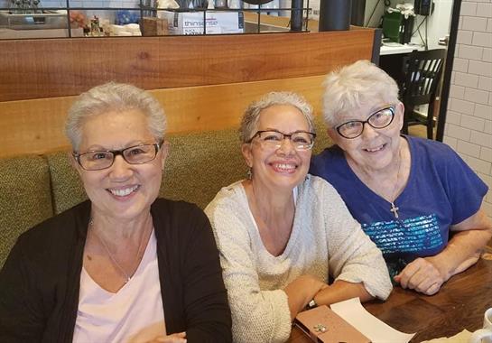 Three women smile brightly while seated at a cafe table, enjoying their time together.