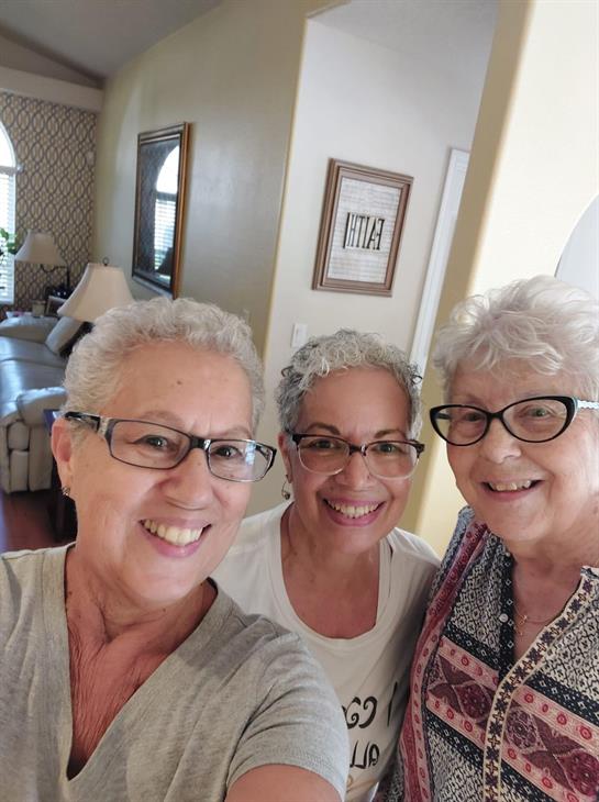Three friends with gray hair smile brightly while posing together in a comfortable living space.