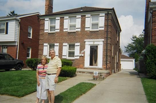 A couple stands happily in front of their brick house, enjoying a sunny day in the suburbs.