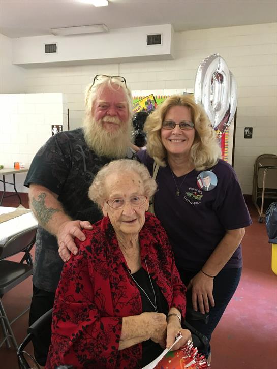 Group of three people gathers around an elderly woman in a red jacket, smiling together warmly.