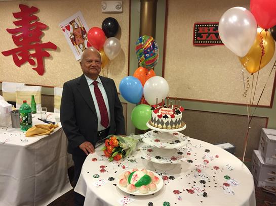 A distinguished man stands proudly by a table adorned with balloons and cake for a celebration.