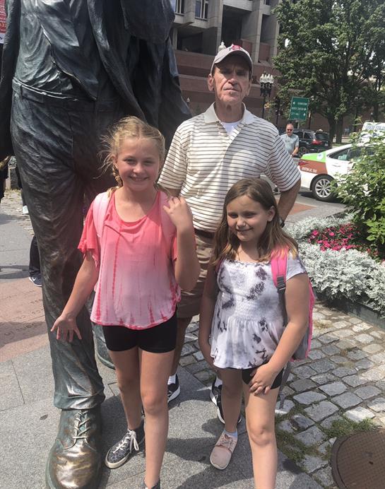 Children smile while standing next to a statue, enjoying a sunny day in a park setting.