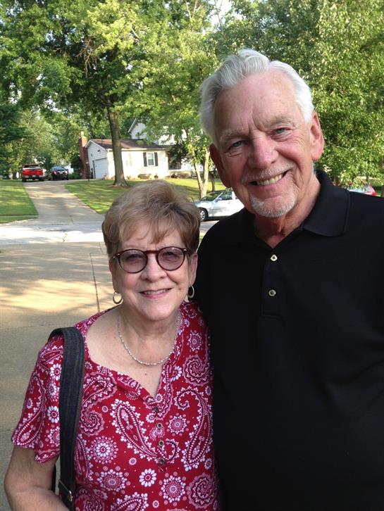 An older couple stands smiling together outside in a sunny suburban area surrounded by greenery.