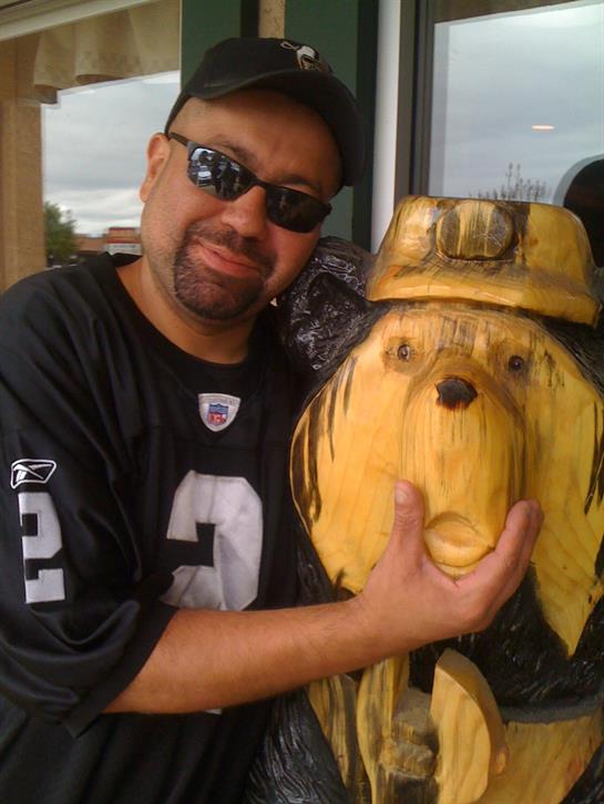 A man smiles and poses with a wooden dog statue dressed in a hat at a storefront.