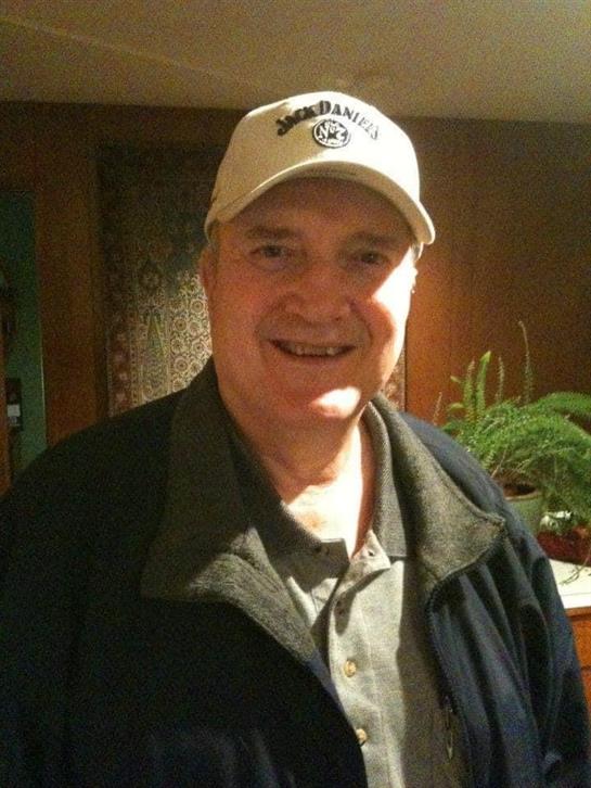 A cheerful man stands indoors, wearing a cap and jacket, while smiling at the camera.