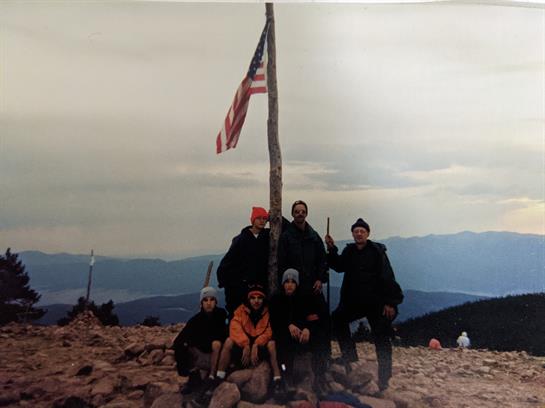 Six climbers pose proudly at the mountain summit under an American flag during their adventure.