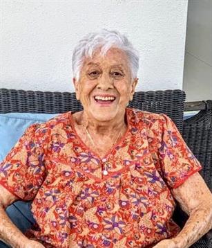 Elderly woman smiles outdoors in a colorful floral shirt, basking in the sunlight.
