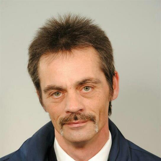 Man with a distinguished mustache stands confidently in front of a light gray backdrop.