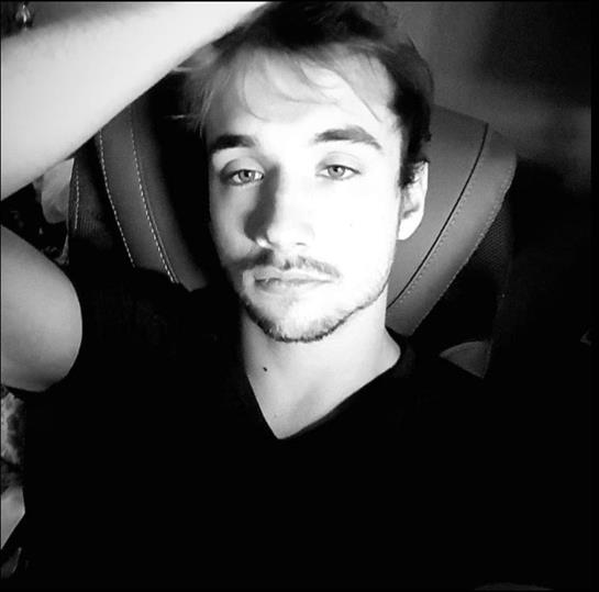 A young man sits back in a chair, deep in thought with a relaxed demeanor in low light.