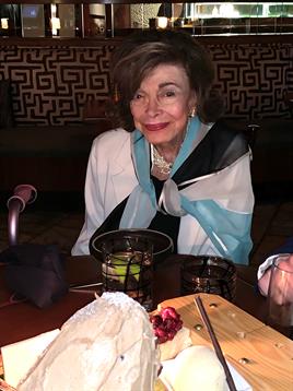 A woman dressed elegantly smiles at the table adorned with a dessert and drinks during dinner.