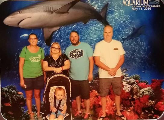 A group of four adults and a child pose together in front of a large shark backdrop at an aquarium.