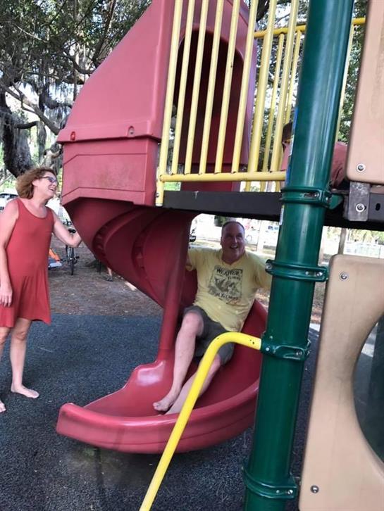 A woman in a red dress playfully interacts with a man sliding down a playground structure.