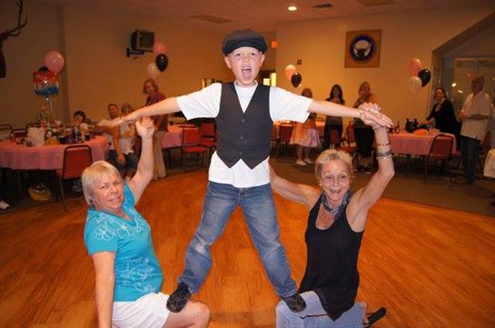 A young boy balances confidently on two women's hands in a festive community hall.