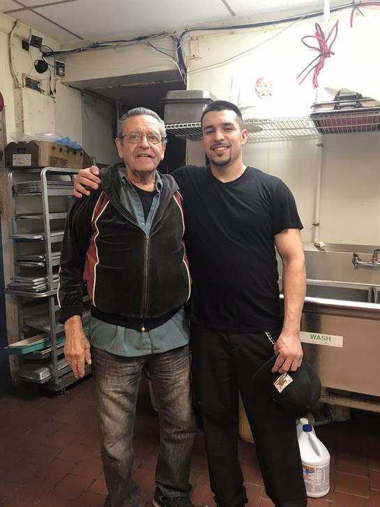 Two men smile in a kitchen, symbolizing teamwork and friendship in a busy setting.