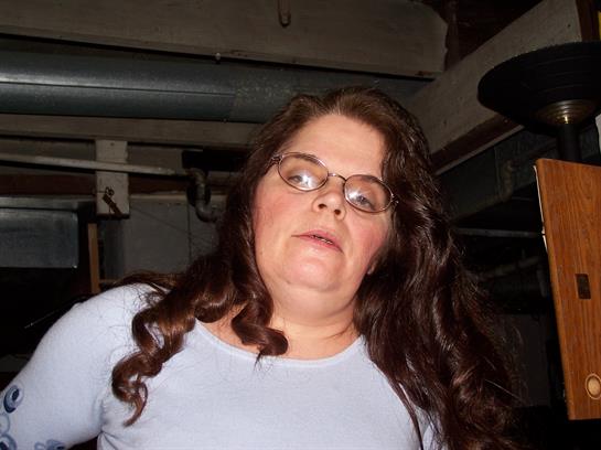 A woman with long curly hair and glasses stands confidently indoors, surrounded by shadows.