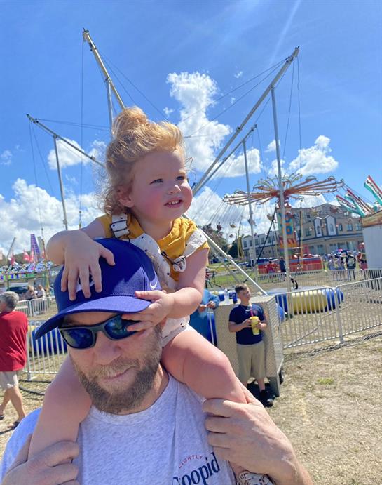 Father lifts his delighted daughter on his shoulders at a bustling fair with rides and activities.