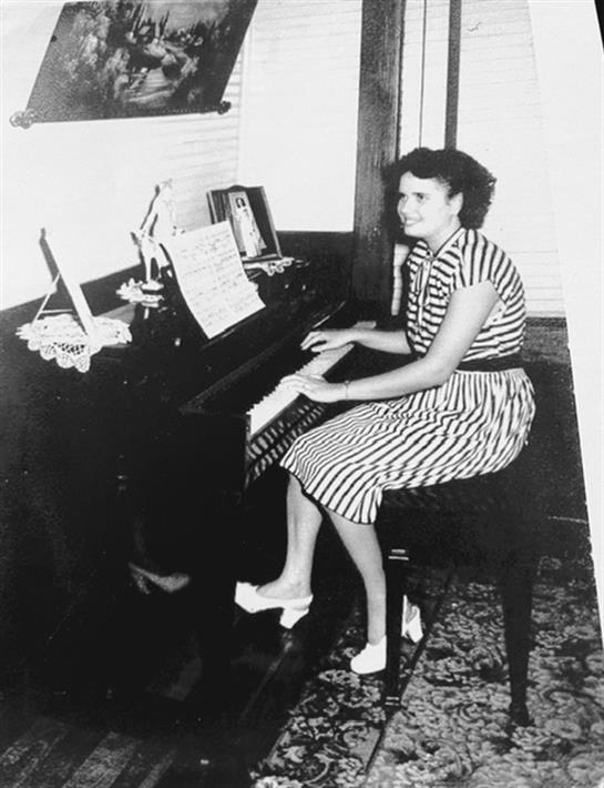 A woman in a striped dress plays a piano in a cozy, vintage room filled with personal items.