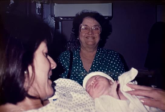 A smiling woman looks on as another woman holds a newborn baby in a cozy hospital room.