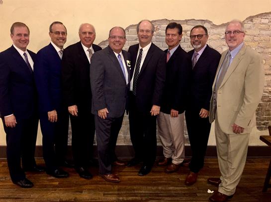 A group of men in suits stands together, smiling for a photo at a formal event indoors.