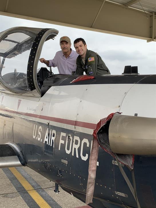 Two individuals are smiling and posing at the cockpit of a U.S. Air Force aircraft in a hangar.