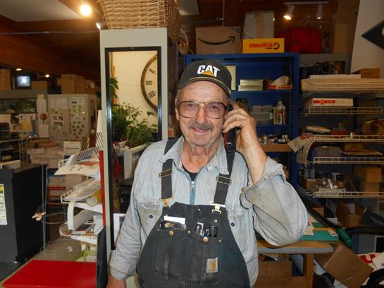 An older mechanic wearing overalls and a cap smiles while using his phone in the workshop.